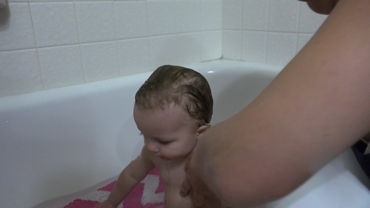 At 0:01 in Westminster, Colorado, the baby looks downward in the bathtub as an adult arm fills the right foreground and white bathroom tile sits behind the tub.
