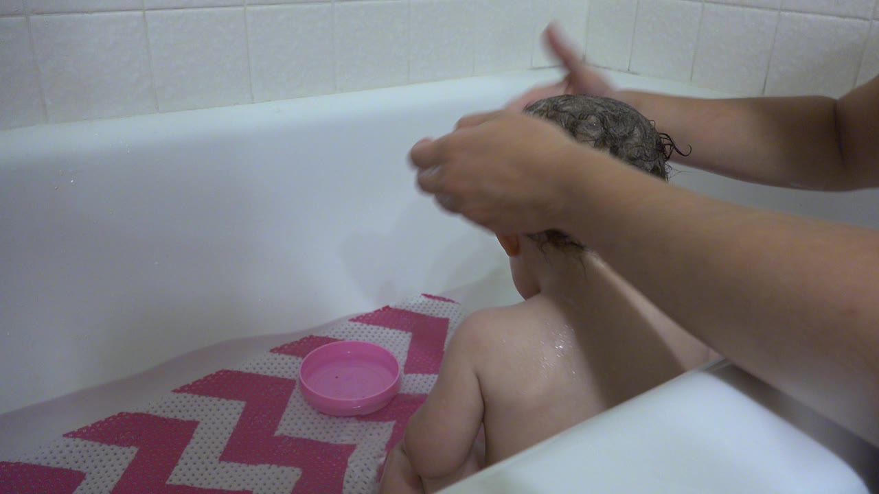 In Westminster, Colorado, adult hands rub soap into a baby's wet hair as the child sits facing away in a white bathtub with tile behind.