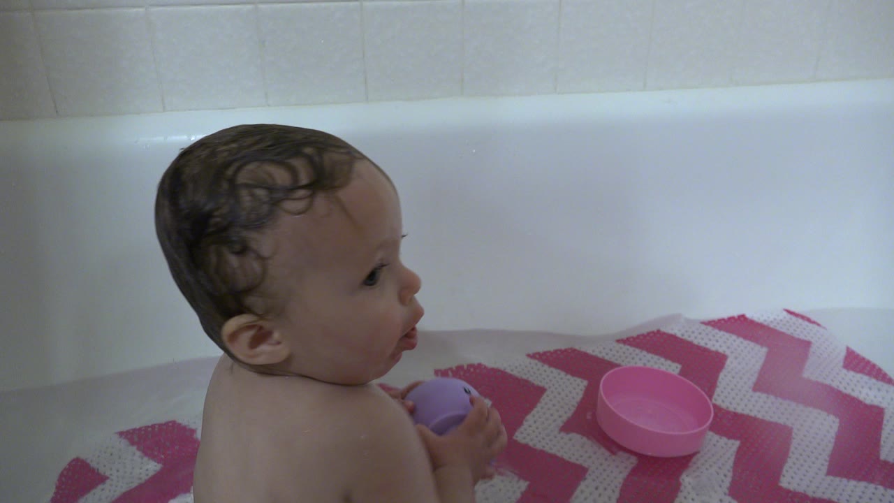 Baby in a white bathtub holding a purple toy beside a pink mat.