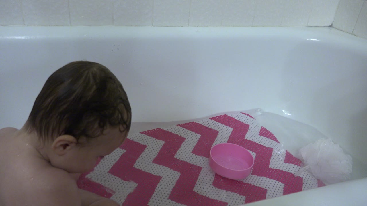 Infant leaning over a pink zigzag bath mat in a white bathtub.