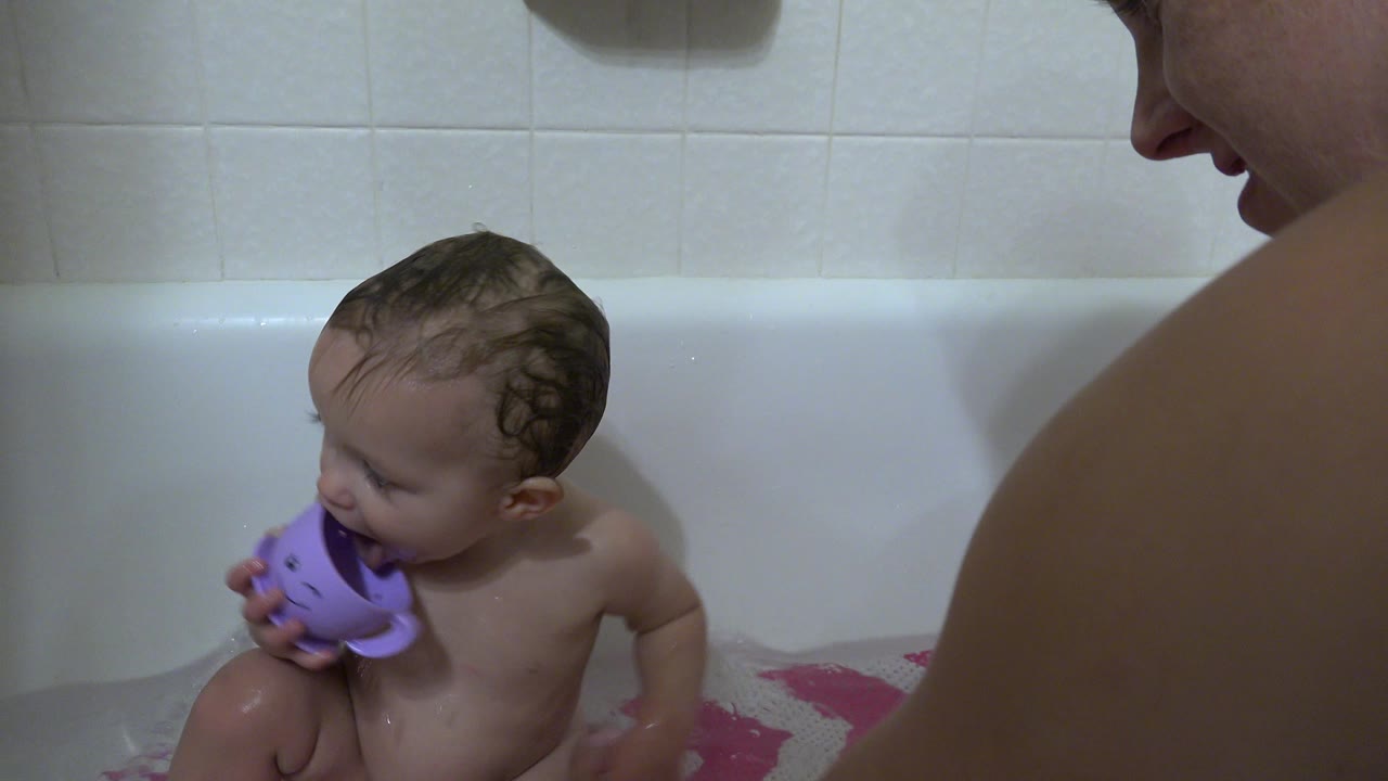 A wet toddler sits in a white bathtub in Westminster, Colorado, holding a purple cup toy while a parent is cropped at the right edge.