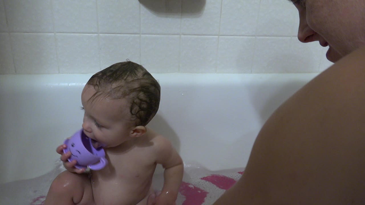 The toddler remains seated in the white tub in Westminster, Colorado, looking down toward the purple cup as the parent's shoulder and face edge are cropped into frame.