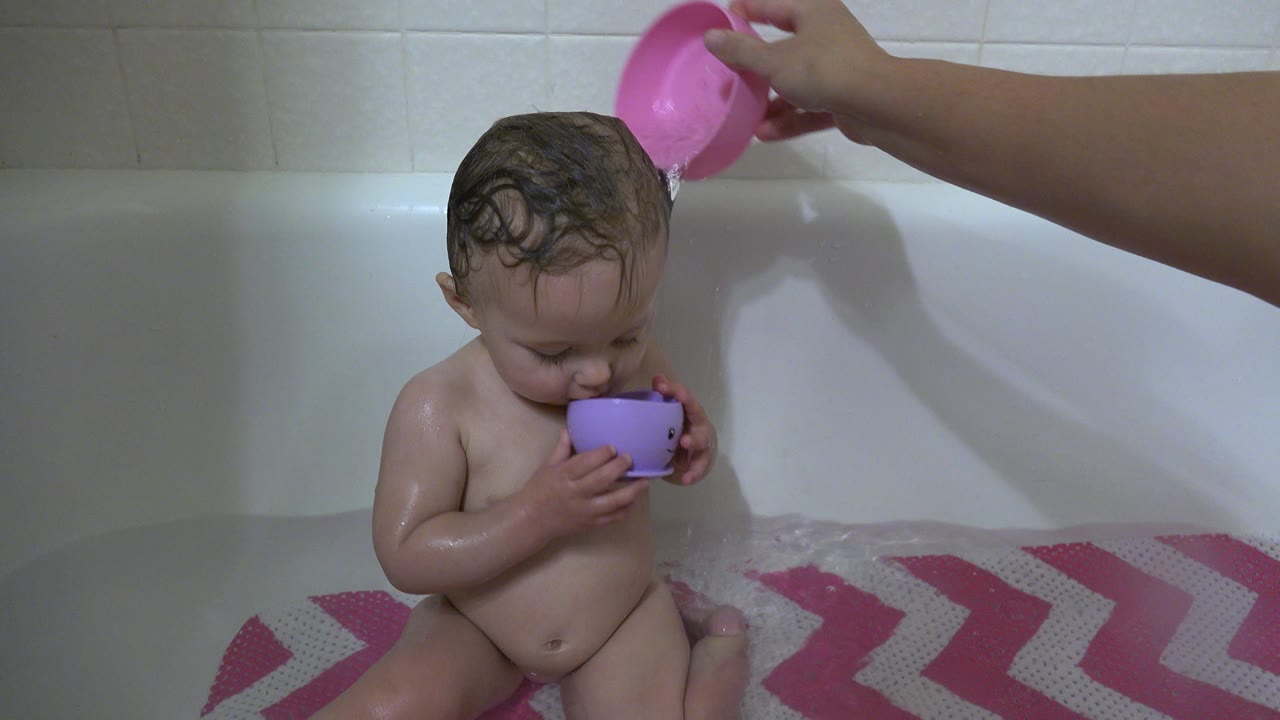 In Westminster, Colorado, the child looks down toward a purple cup while the adult's pink cup remains above the bathtub rim and water ripples across the bath mat.