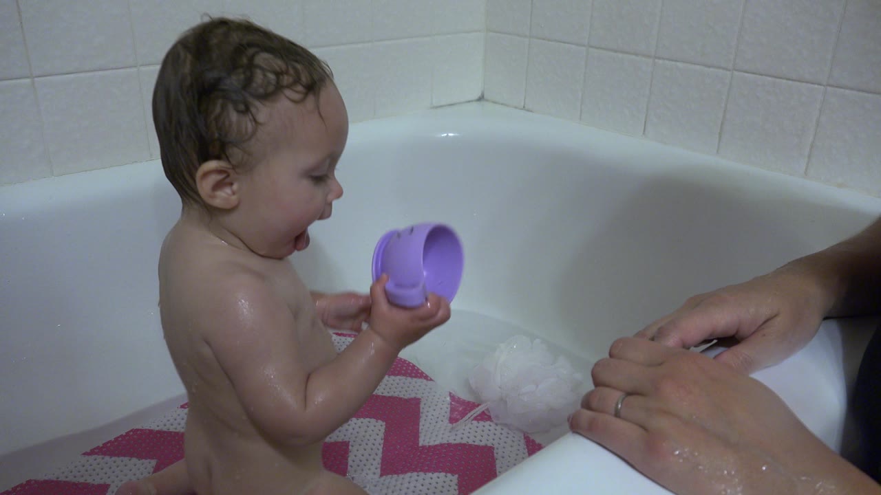 The baby remains seated in the white bathtub in Westminster, Colorado, with wet hair, a purple cup in both hands, and adult hands visible on the right edge.