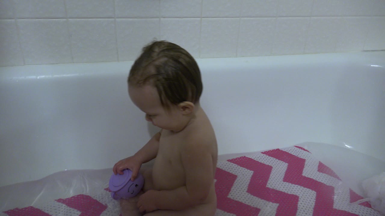 A baby in Westminster, Colorado sits in a white bathtub looking down at a purple bath toy on a pink-and-white chevron mat.