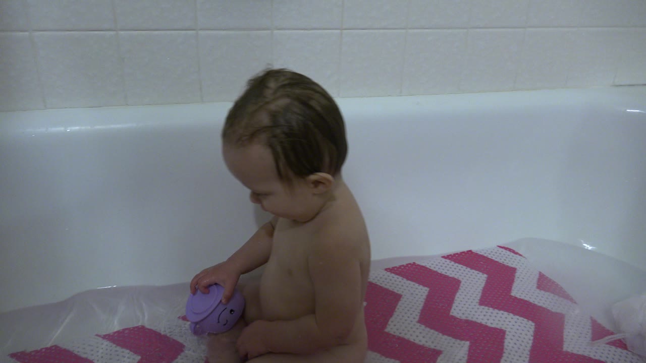 A baby in Westminster, Colorado remains seated in the tub, head tilted down toward the purple toy near the bath mat.