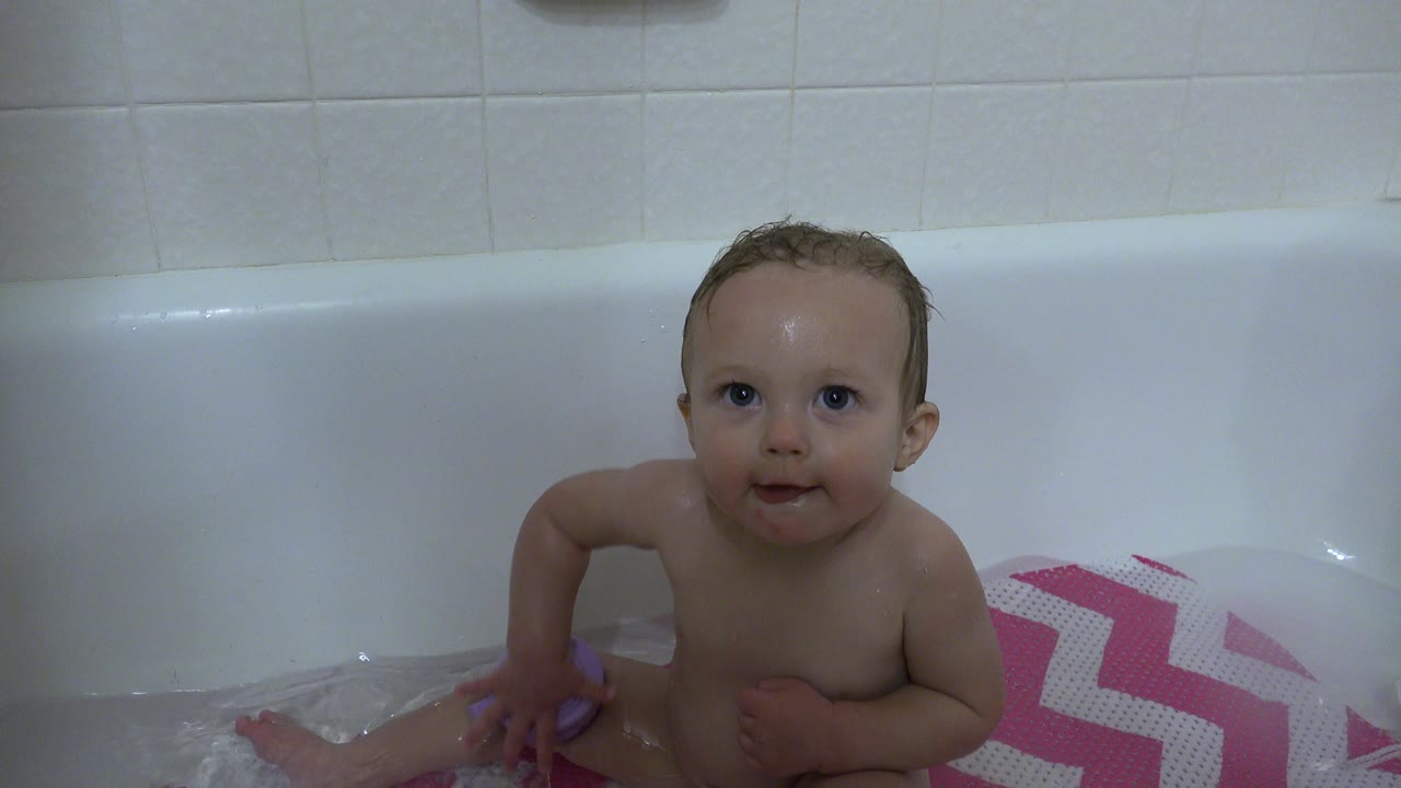 Baby seated in a white bathtub in Westminster, Colorado, facing the camera with shallow bathwater and a pink chevron mat visible at the side.
