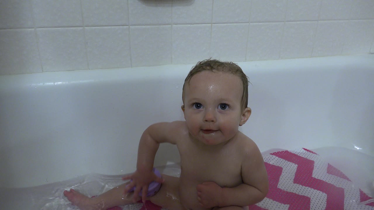 Baby in a Westminster, Colorado bathtub looks toward the camera while one arm rests near the water and the pink chevron mat remains at frame right.