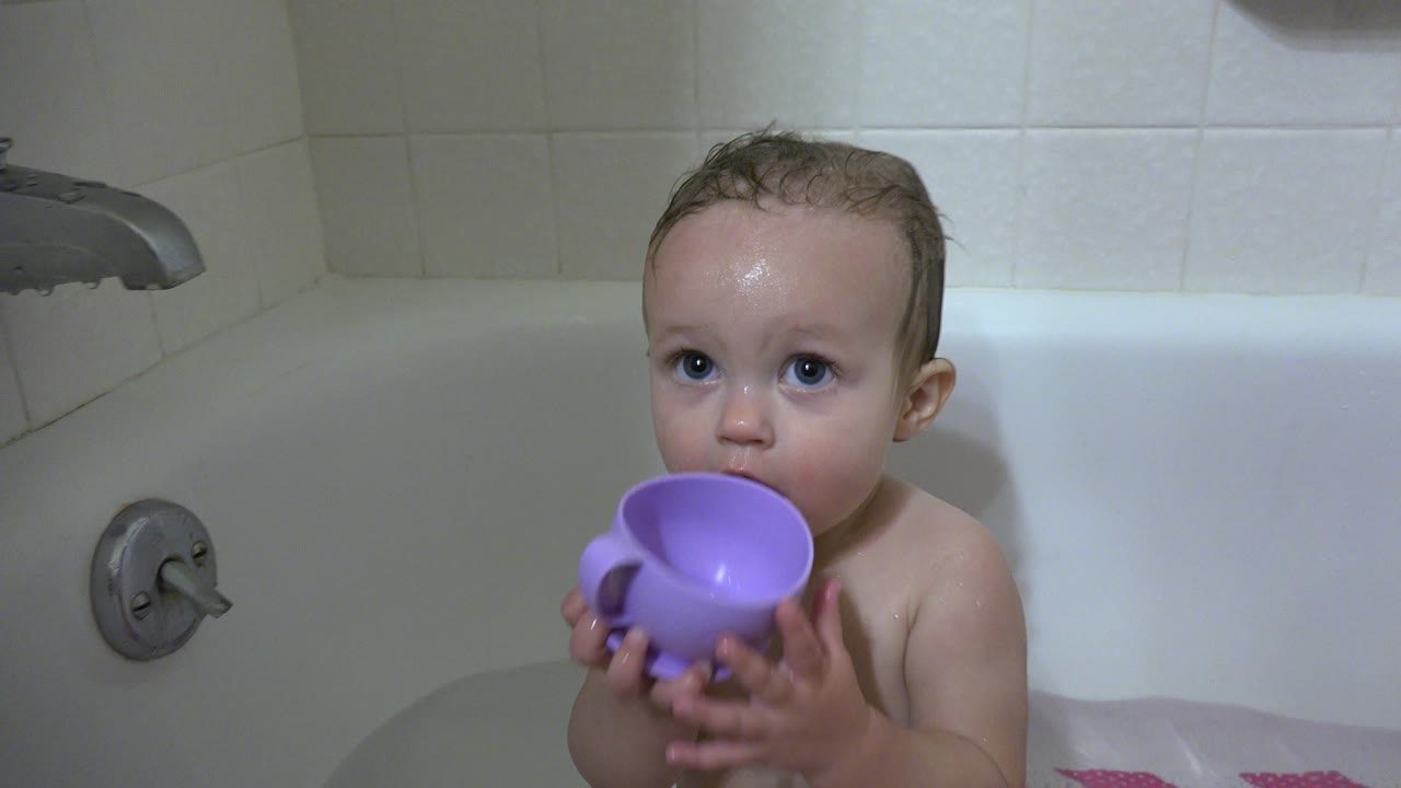 Baby in a bathtub holding a purple mug near their mouth.