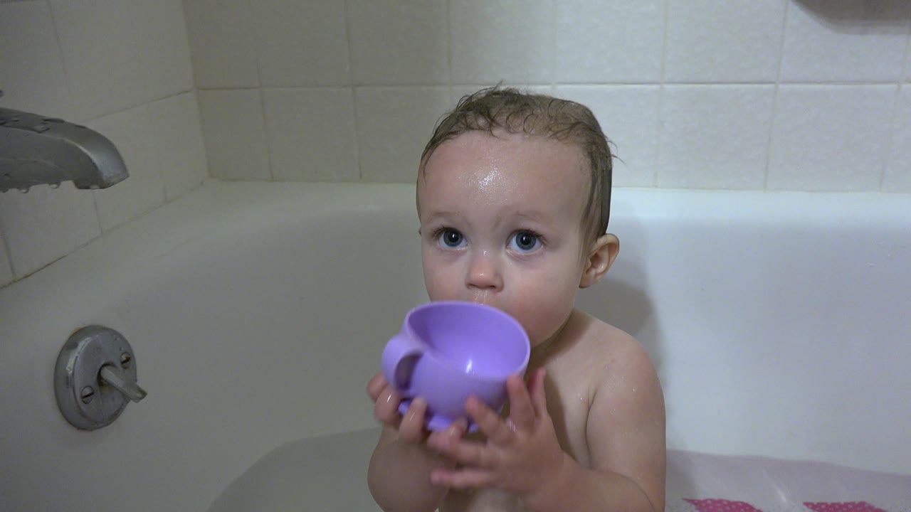 In Westminster, Colorado, the baby remains centered in the bathtub with both hands around the purple mug and bath fixtures visible behind them.