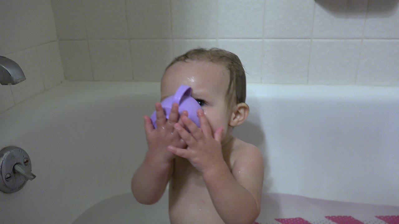 A wet baby in a white bathtub in Westminster, Colorado holds a purple cup against their face near a silver tub handle.