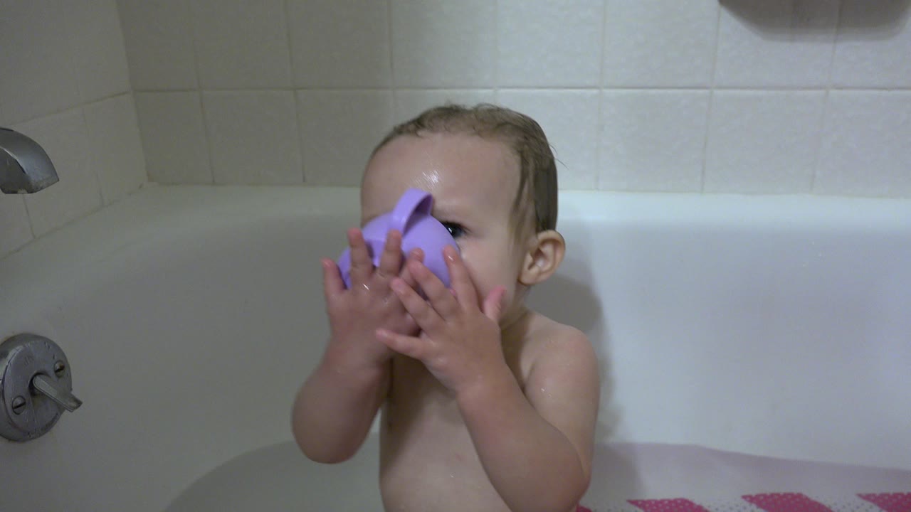 The baby remains seated in the Westminster, Colorado bathtub with both hands covering a purple cup held over the face.