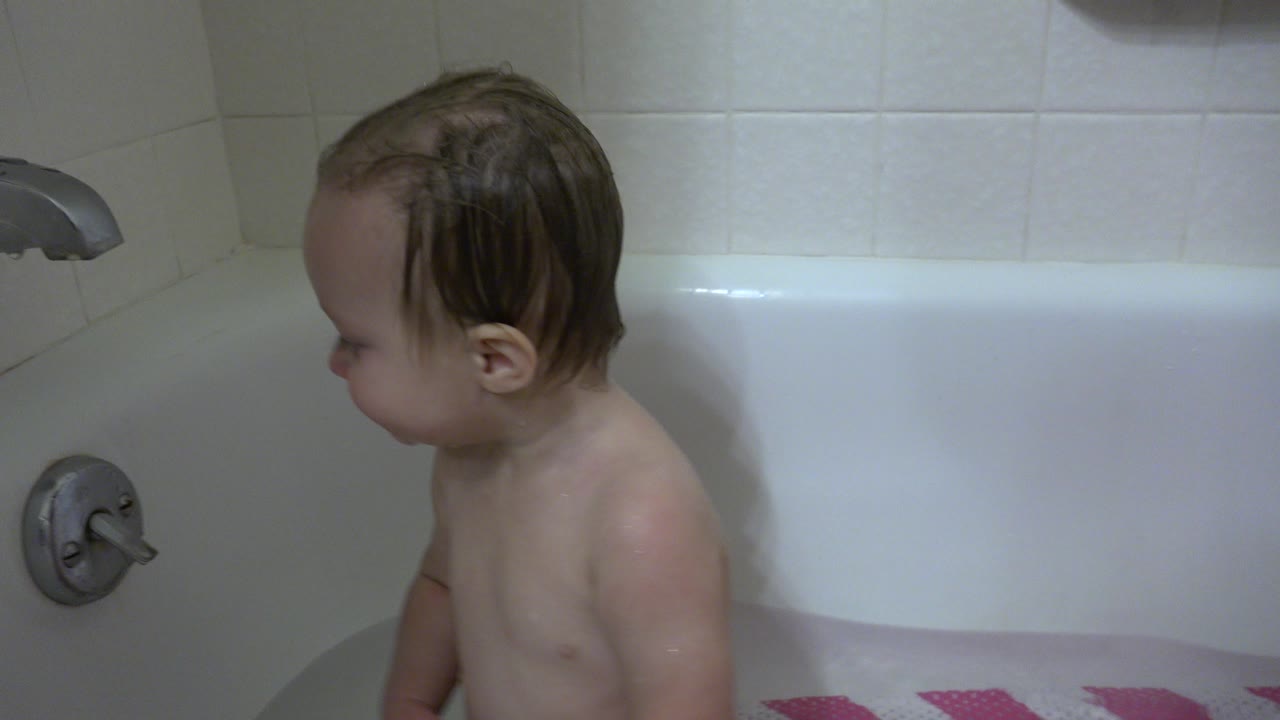 Wet baby sitting in a bathtub beside a faucet.