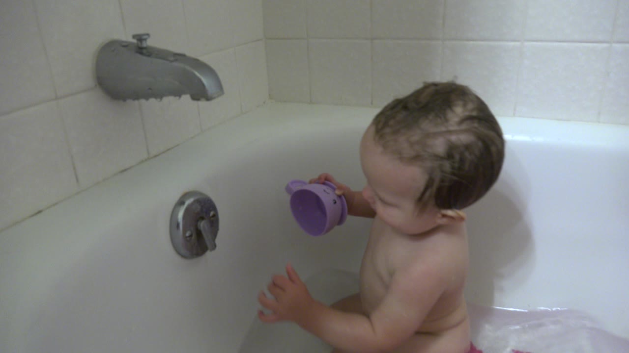Baby sitting in a bathtub holding a purple toy cup.