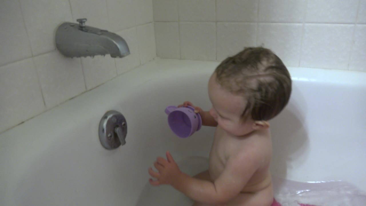 In Westminster, Colorado, the baby remains seated in the bathtub with the purple toy cup raised, facing the chrome control on the tub wall.