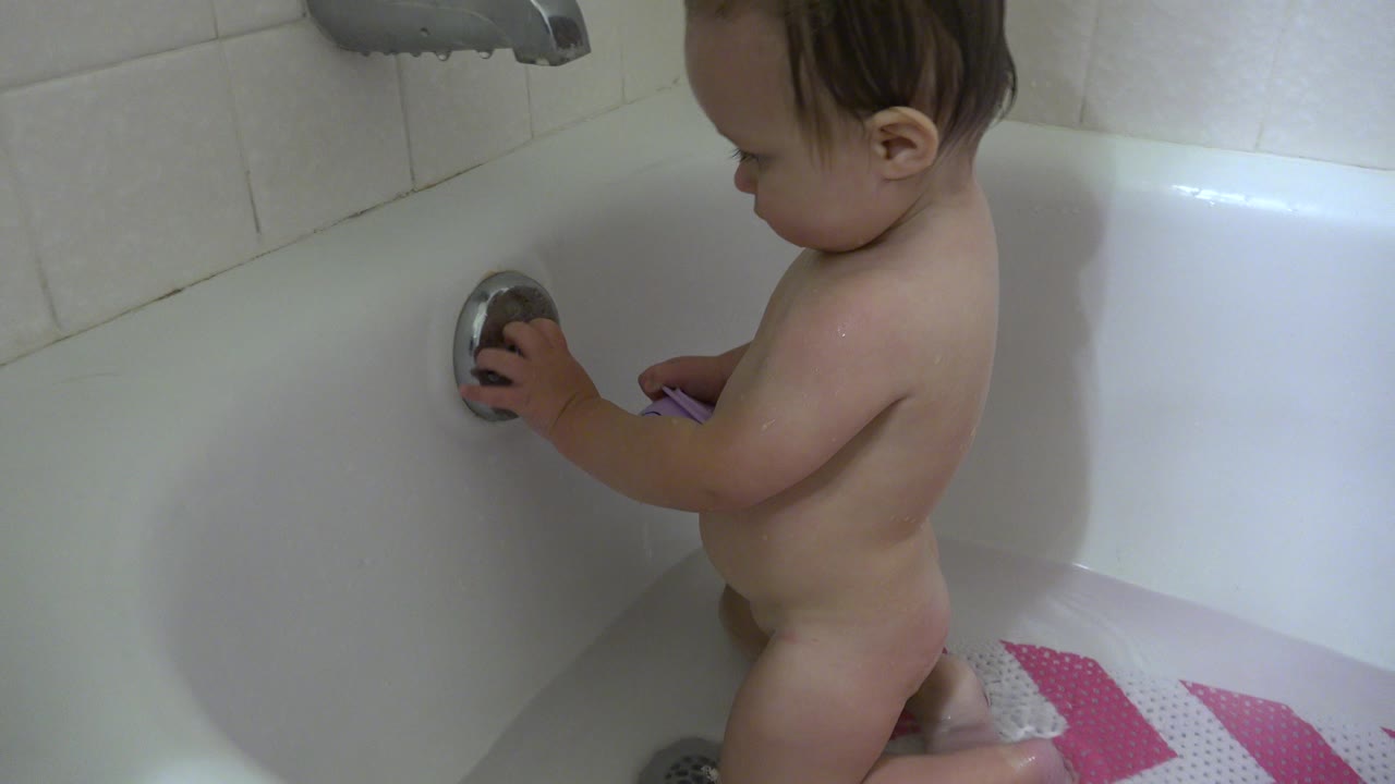 Baby standing in a white bathtub touching the chrome drain cover.