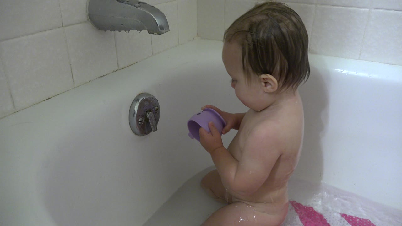 Young child seated in a bathtub holding a purple cup near a chrome handle.