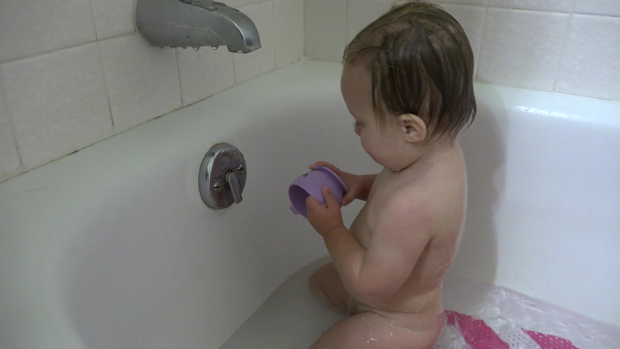 Young child seated in the Westminster bathtub looks down while handling the purple cup near the tub wall and metal control.