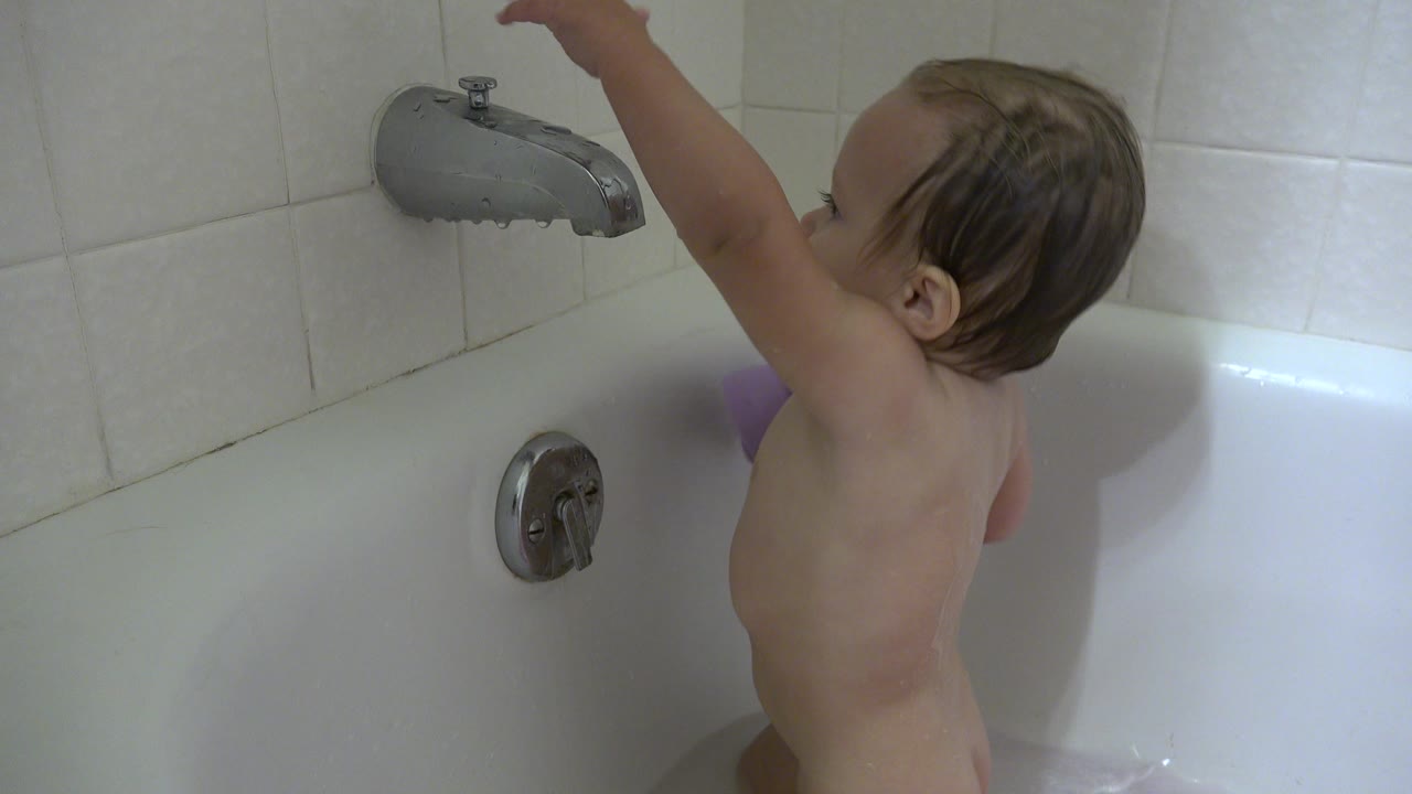 A wet-haired baby in a white bathtub in Westminster, Colorado reaches one arm up toward the chrome faucet on a tiled wall.