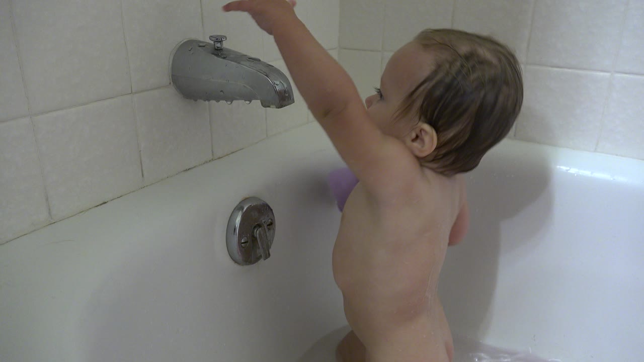 The baby stands in the white tub with one arm lifted toward the faucet, with white wall tile and the chrome control visible beside the child.