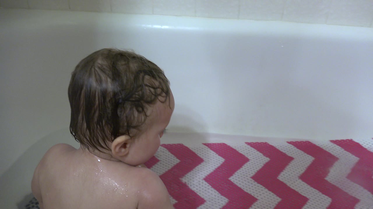 At 0:00 in Westminster, Colorado, a wet toddler sits with their back to camera in a white bathtub beside a pink and white chevron bath mat.