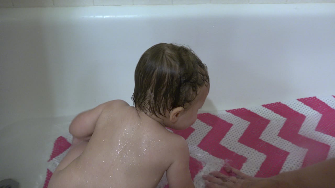 Toddler leans forward on the pink and white mat in the bathtub.