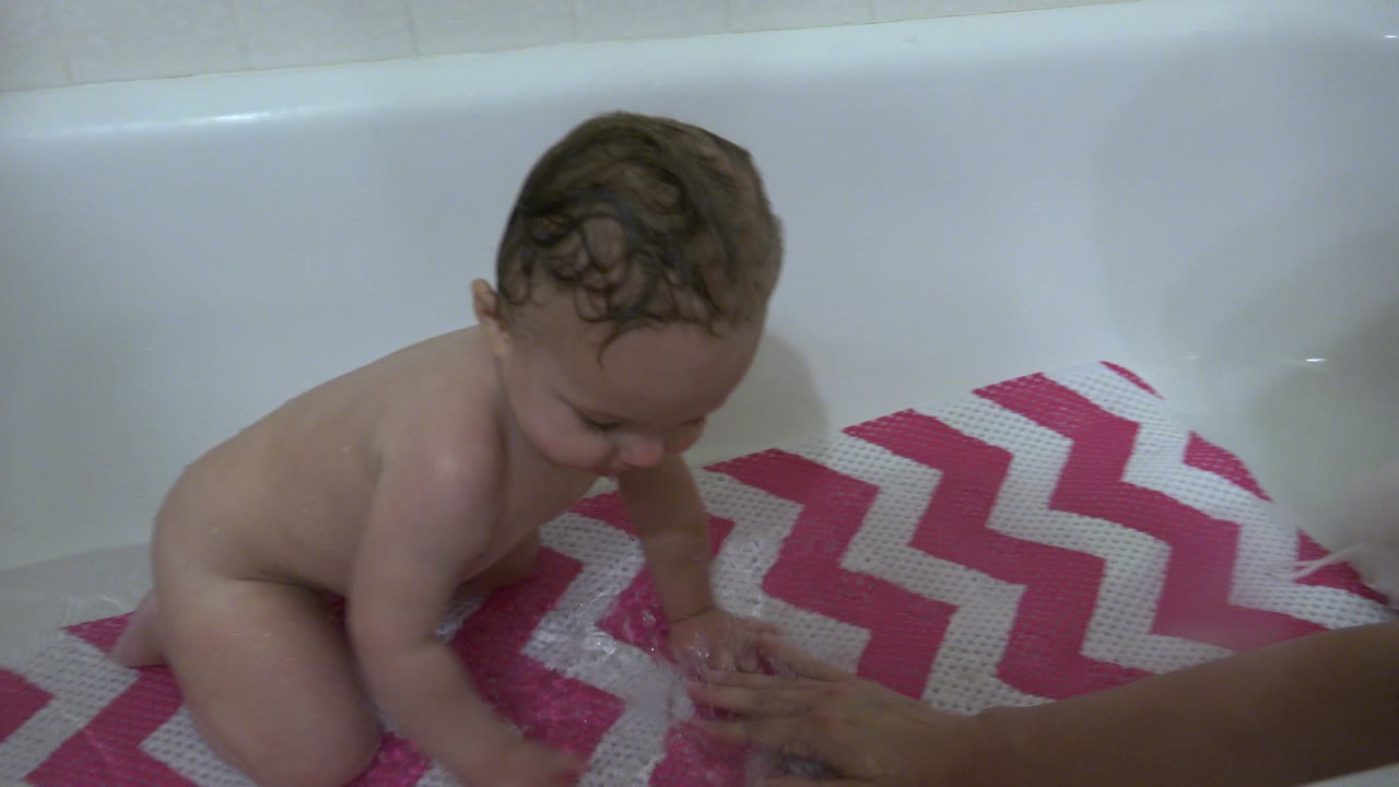 Baby sitting in a bathtub beside a pink chevron bath mat and an adult hand.