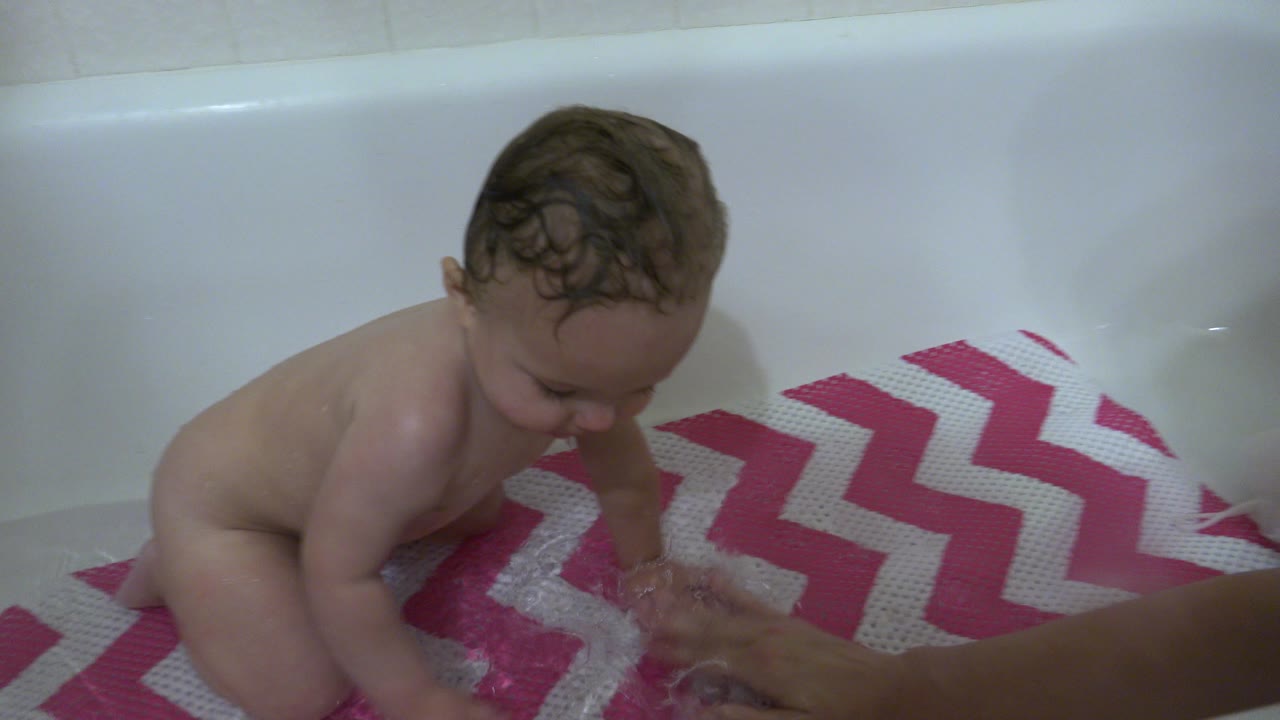 At 0:01 in Westminster, Colorado, the baby leans forward over shallow bath water while an adult hand is visible near the patterned mat.
