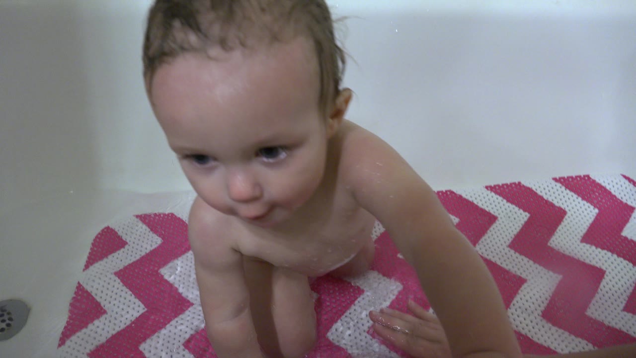 The baby remains close to the camera in the white bathtub, leaning forward over the chevron bath mat with a partial adult hand entering from below.