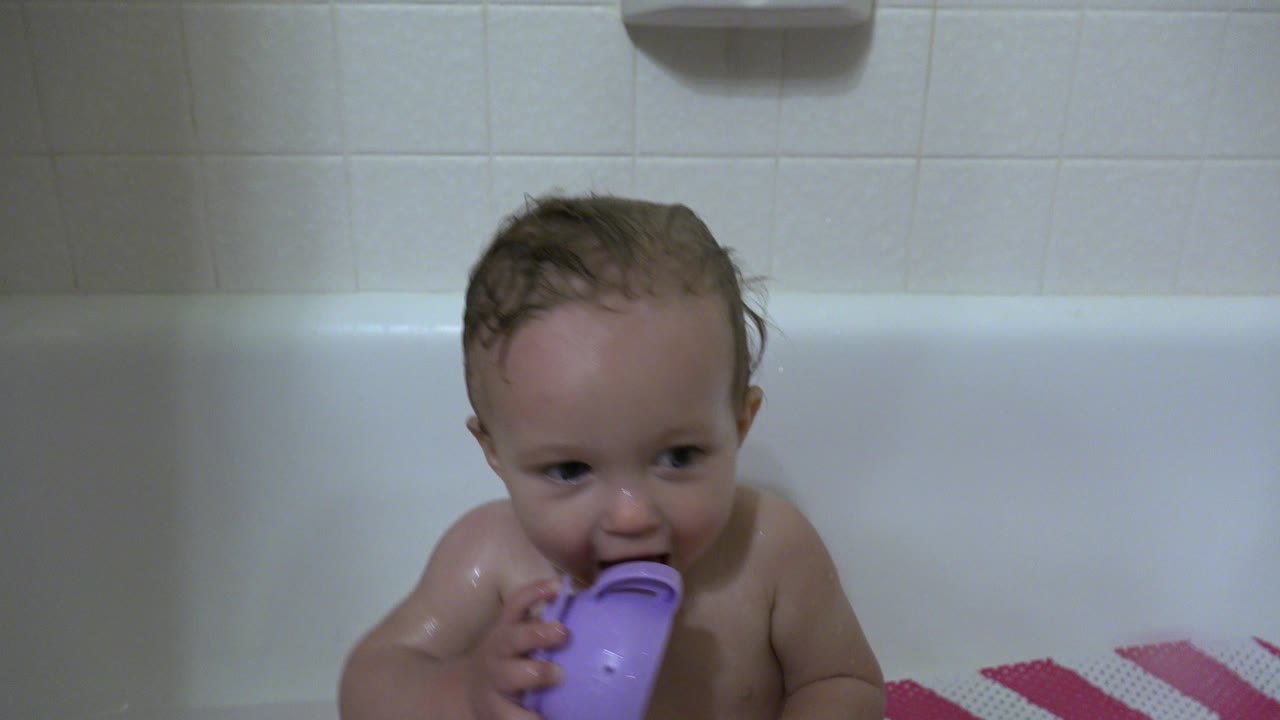 Wet baby sitting in a white bathtub holding a purple cup.