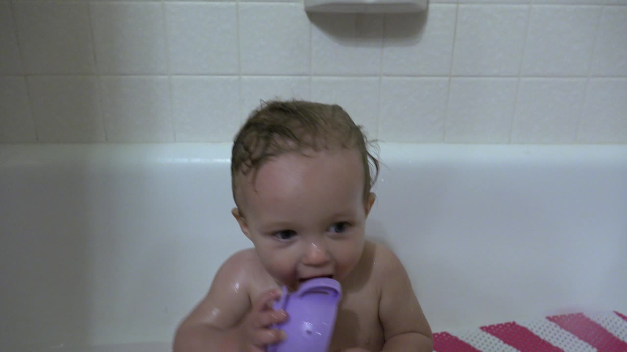 The baby remains seated in the white tub in Westminster, Colorado, with wet hair and the purple cup lifted close to the face.