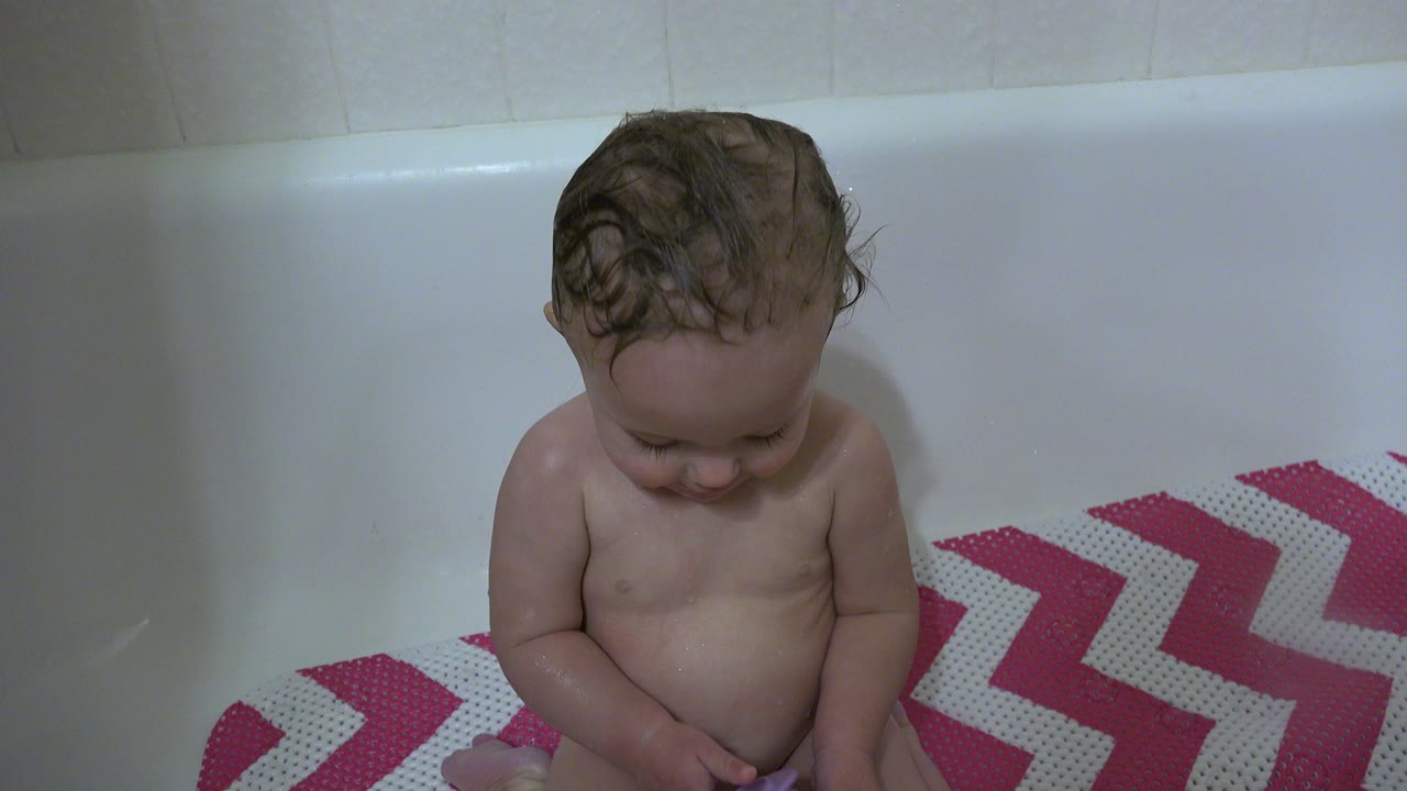 Baby sitting in a white bathtub looking down.