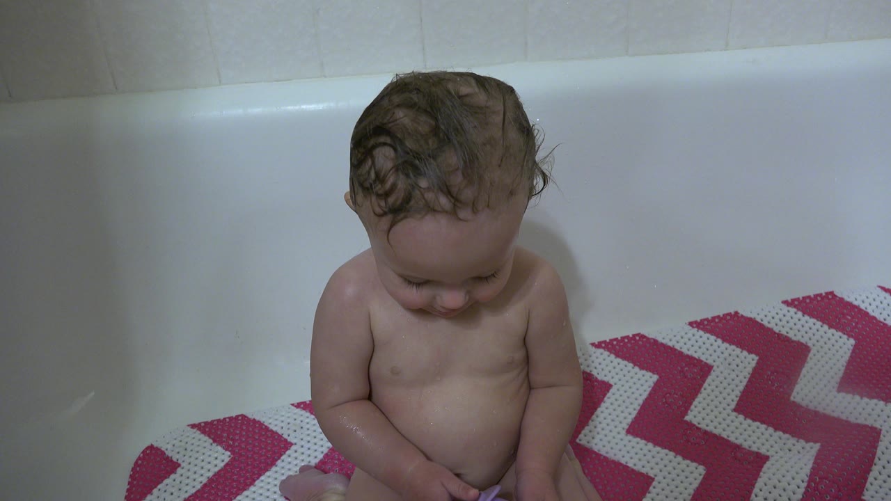 The baby remains seated in the white tub in Westminster, Colorado, with wet hair, head lowered, and the chevron bath mat visible to the right.