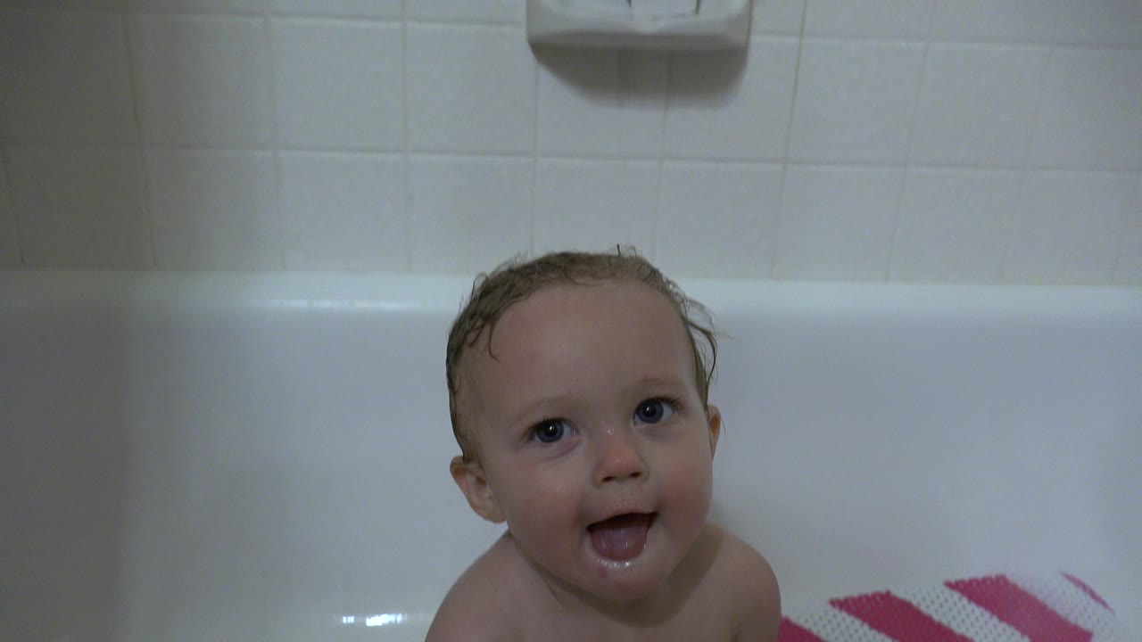 A wet baby looks up with an open mouth from a white bathtub in Westminster, Colorado, with pale tile and part of a pink chevron bath mat visible.