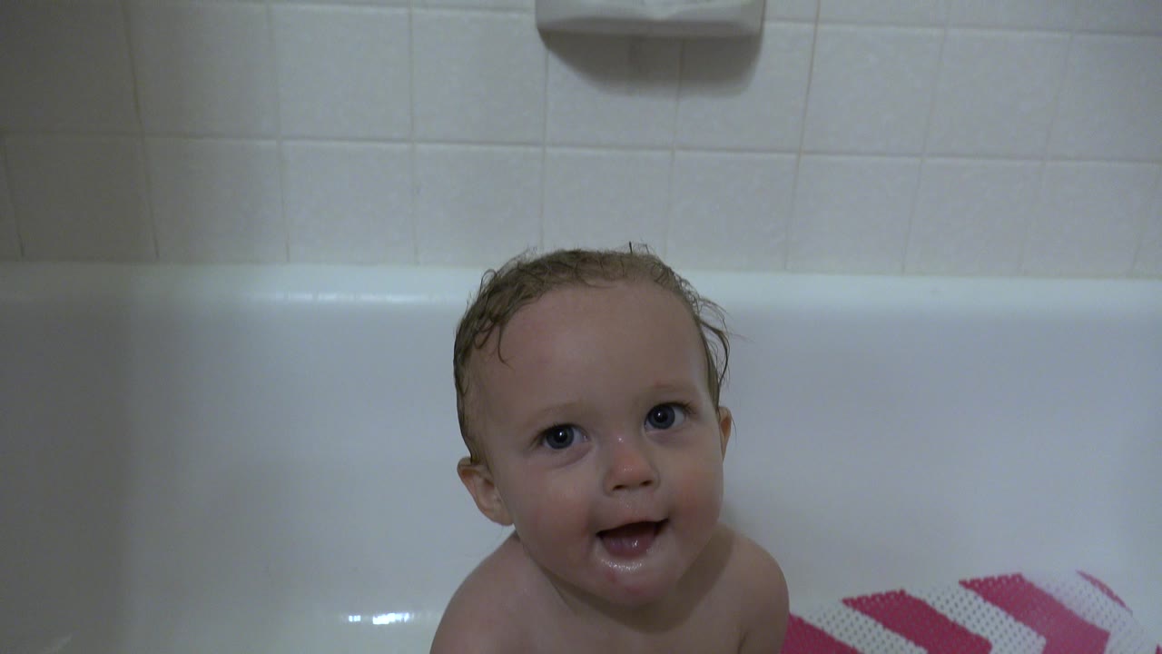 The baby remains near the center of the white tub in Westminster, Colorado, facing the camera with wet hair and the chevron bath mat along the lower right edge.
