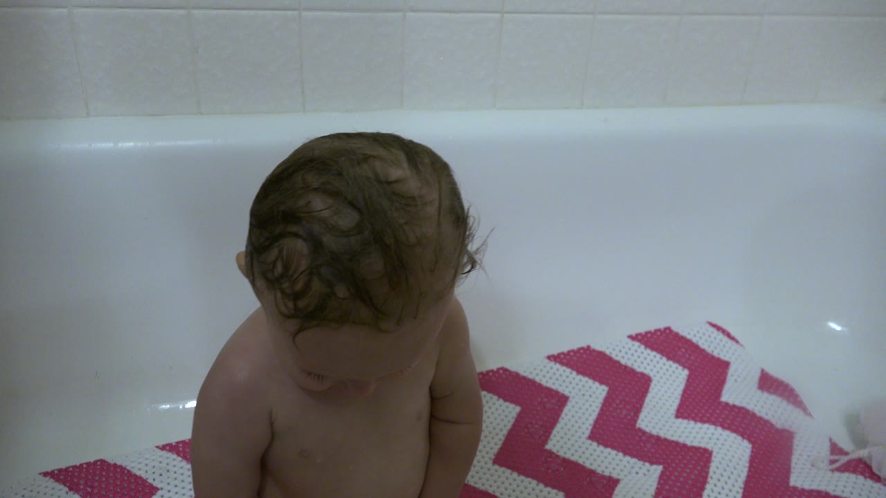 Wet-haired baby looking down in a white bathtub.