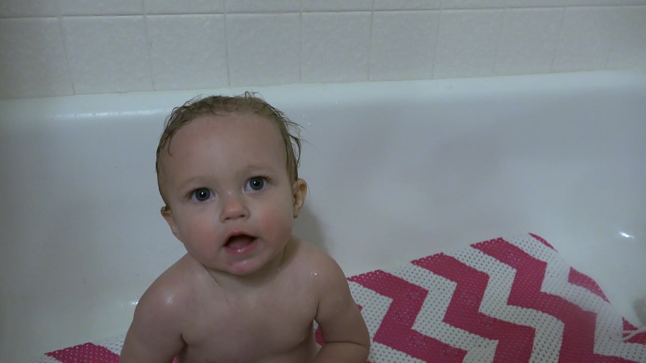 Wet baby in Westminster, Colorado sits in a white bathtub, looking toward the camera with pale tile behind and a pink chevron bath mat at the side.