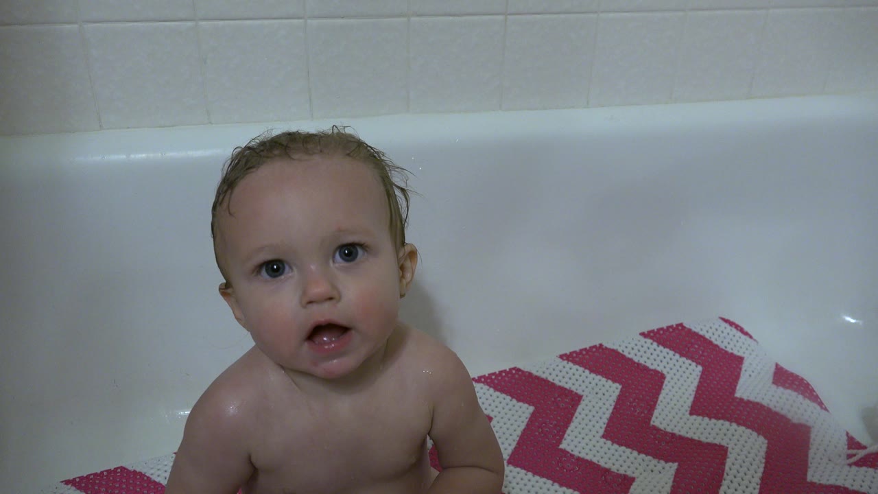 Baby in a white bathtub in Westminster, Colorado faces the camera, with wet hair, pale bathroom tile, and a pink-and-white chevron mat visible to the right.