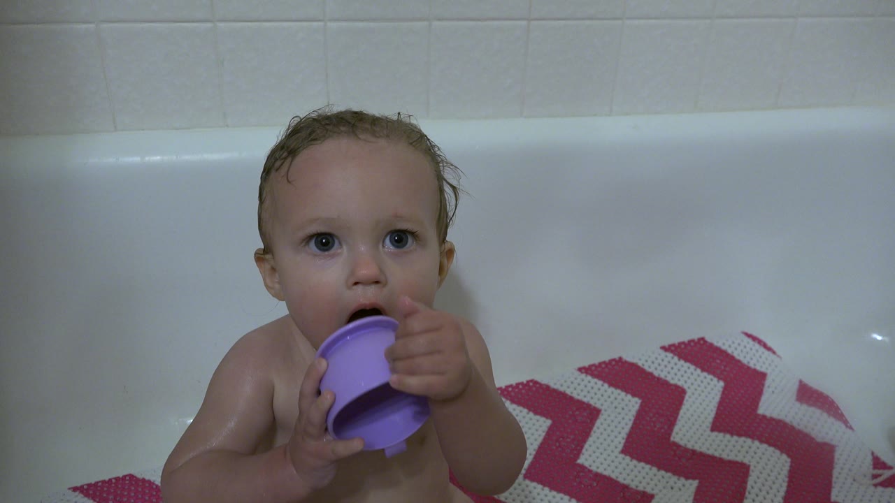 Baby in Westminster, Colorado holds a purple cup with both hands near their mouth while seated in the white bathtub beside the pink chevron mat.