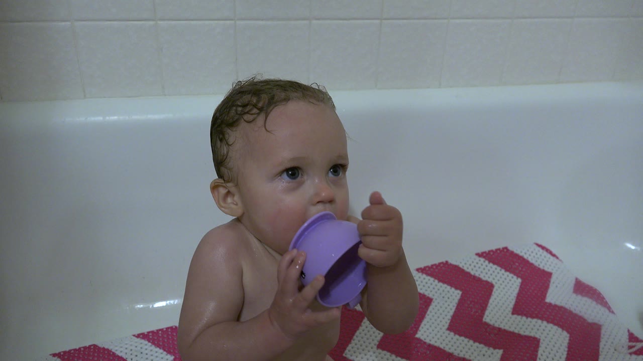 Baby turns sideways while mouthing a purple cup in the bathtub.