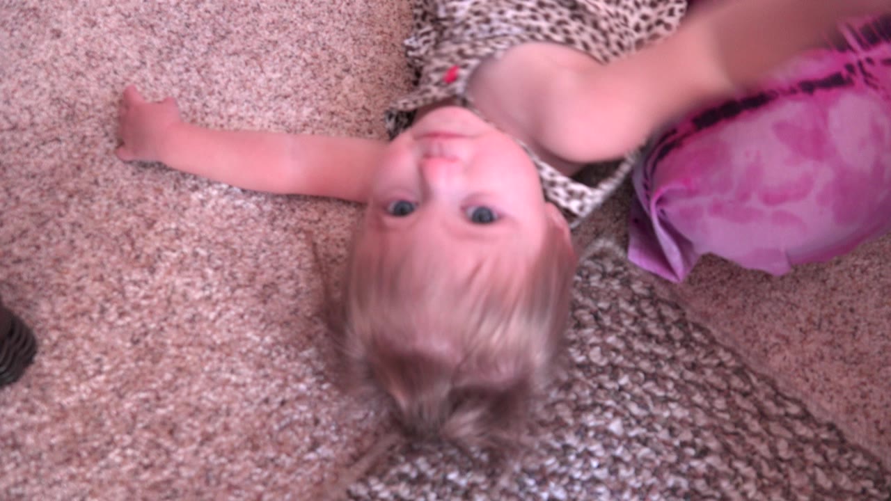 A young girl lies on a beige carpet in Westminster, Colorado, looking up toward the camera beside a pink cushion and textured rug.