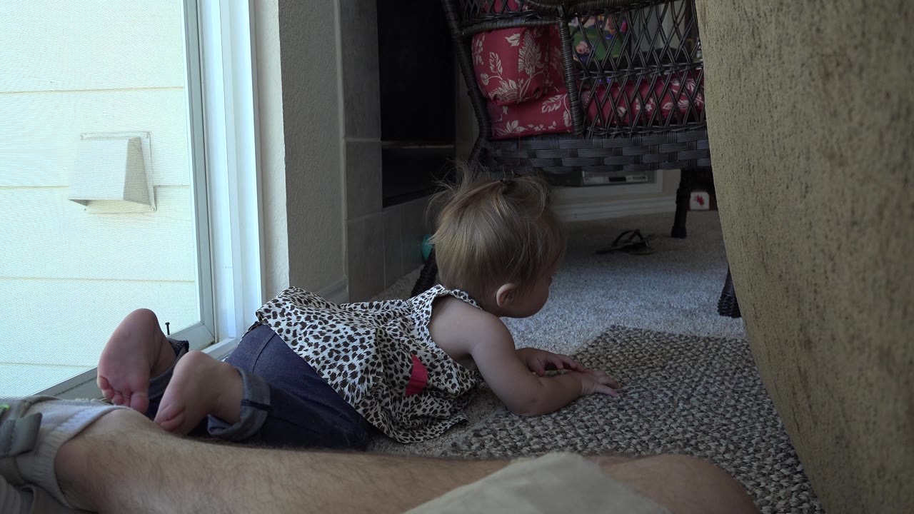 At 0:01 in Westminster, Colorado, the child remains on hands and knees, moving across the carpet with the bright sliding door to the left and a chair with red cushions behind.