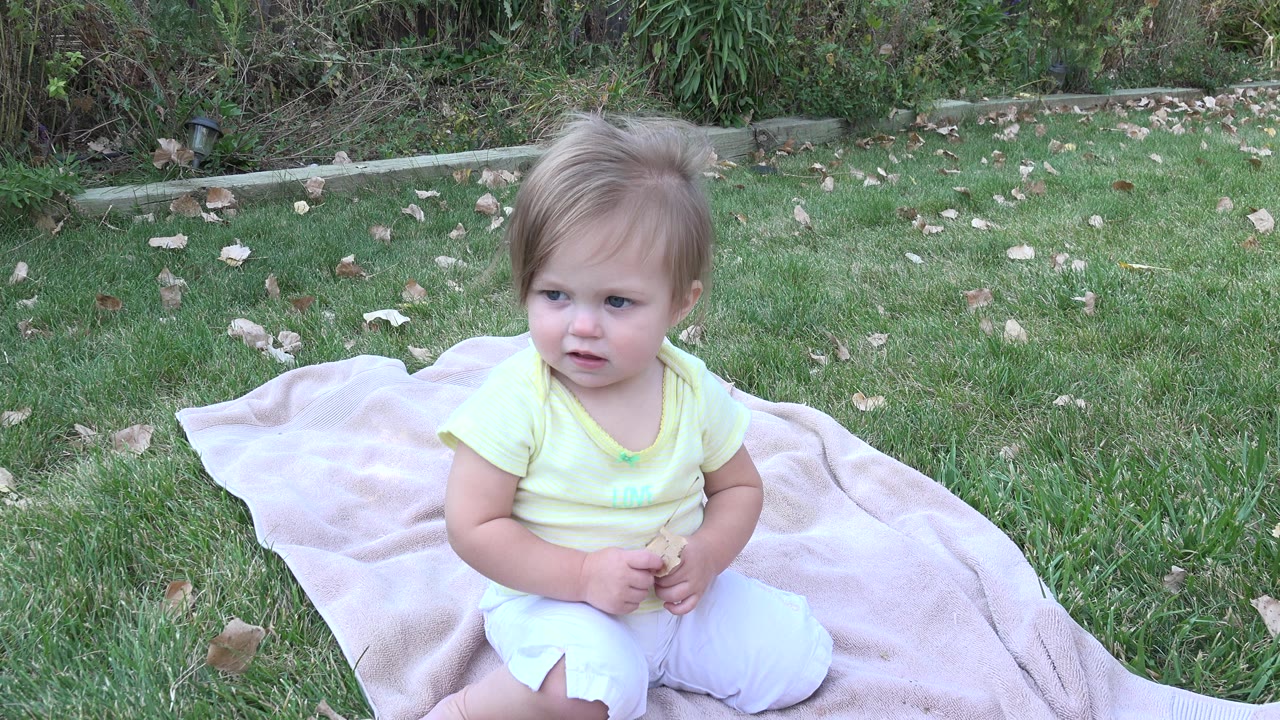 At 0:00, a baby sits on a light pink blanket in a Westminster, Colorado grassy yard, looking off to the side with shrubs behind the lawn.