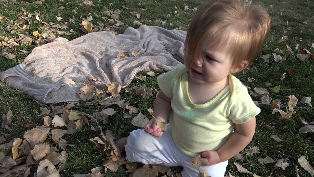 Toddler on grass holding leaves beside a light blanket.