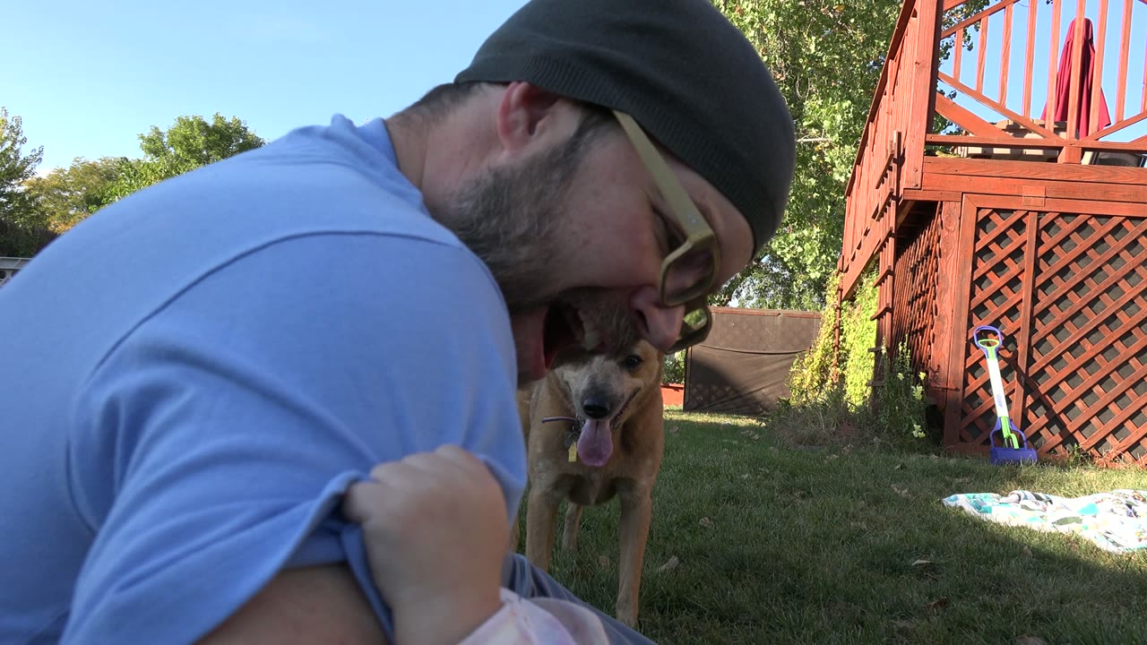 In a Westminster, Colorado backyard, a bearded man in glasses leans close to a small child while a brown dog stands behind them near the lawn and red deck.