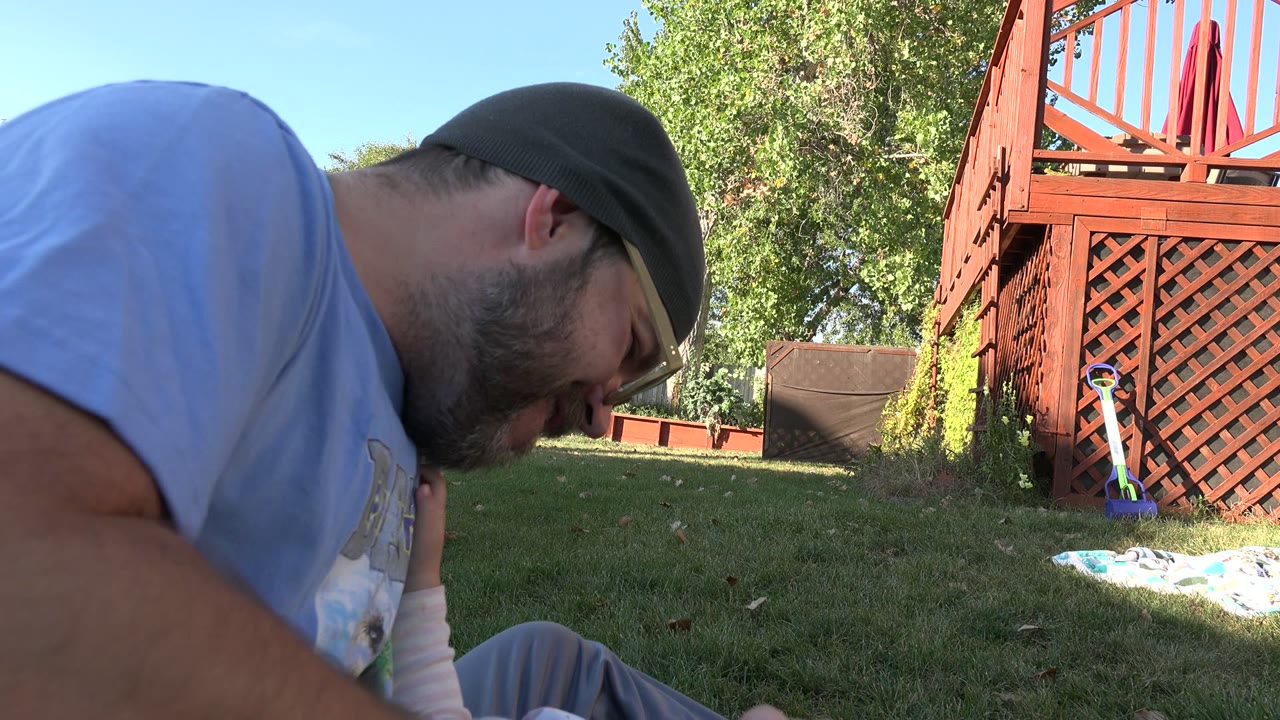Man in glasses leans over a baby on grass beside a backyard deck.