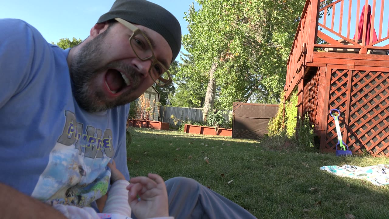In Westminster, Colorado, the man faces the camera with his mouth open while a baby's legs and hand appear in the foreground beside the red deck and trees.