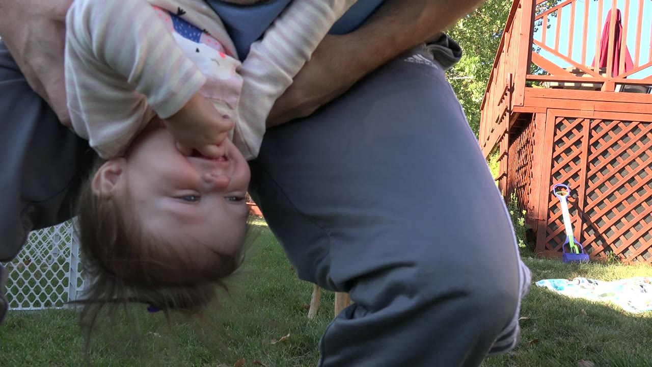 Child held upside down near an adult's legs in a backyard.