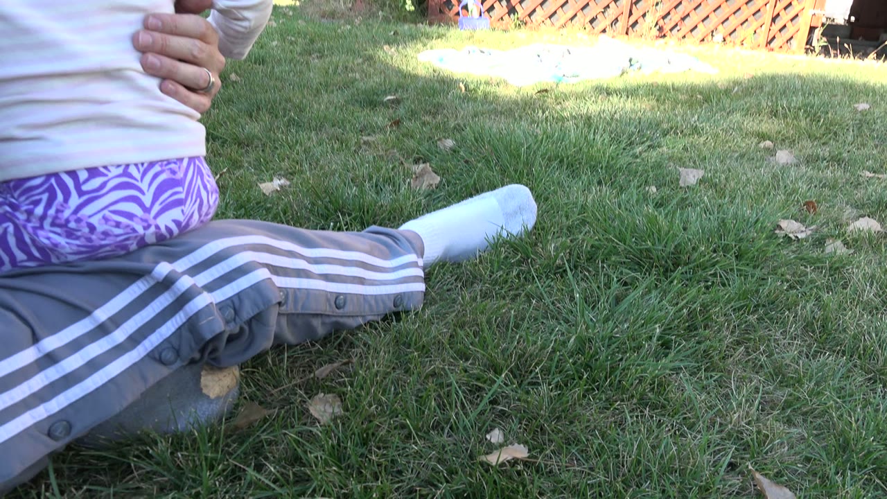 At 0:01 in Westminster, Colorado, a toddler in purple patterned pants sits on an adult's lap beside a stretched socked leg on the grass.