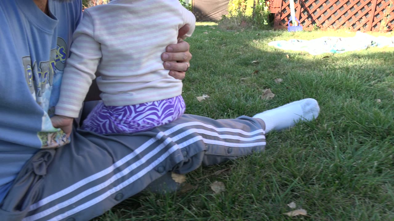 At 0:02 in Westminster, Colorado, a father in a blue shirt holds a toddler close on his lap while sitting in backyard grass near a wooden fence.